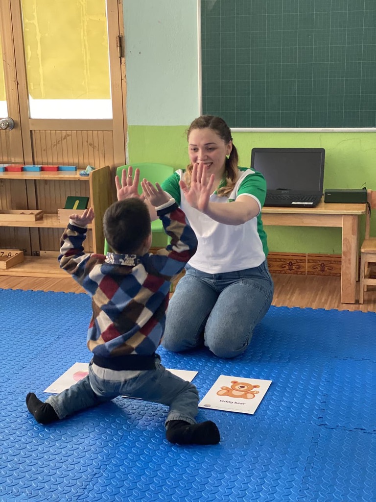 Female English teacher high-fiving toddler on classroom floor with flashcards kindergarten Vietnam