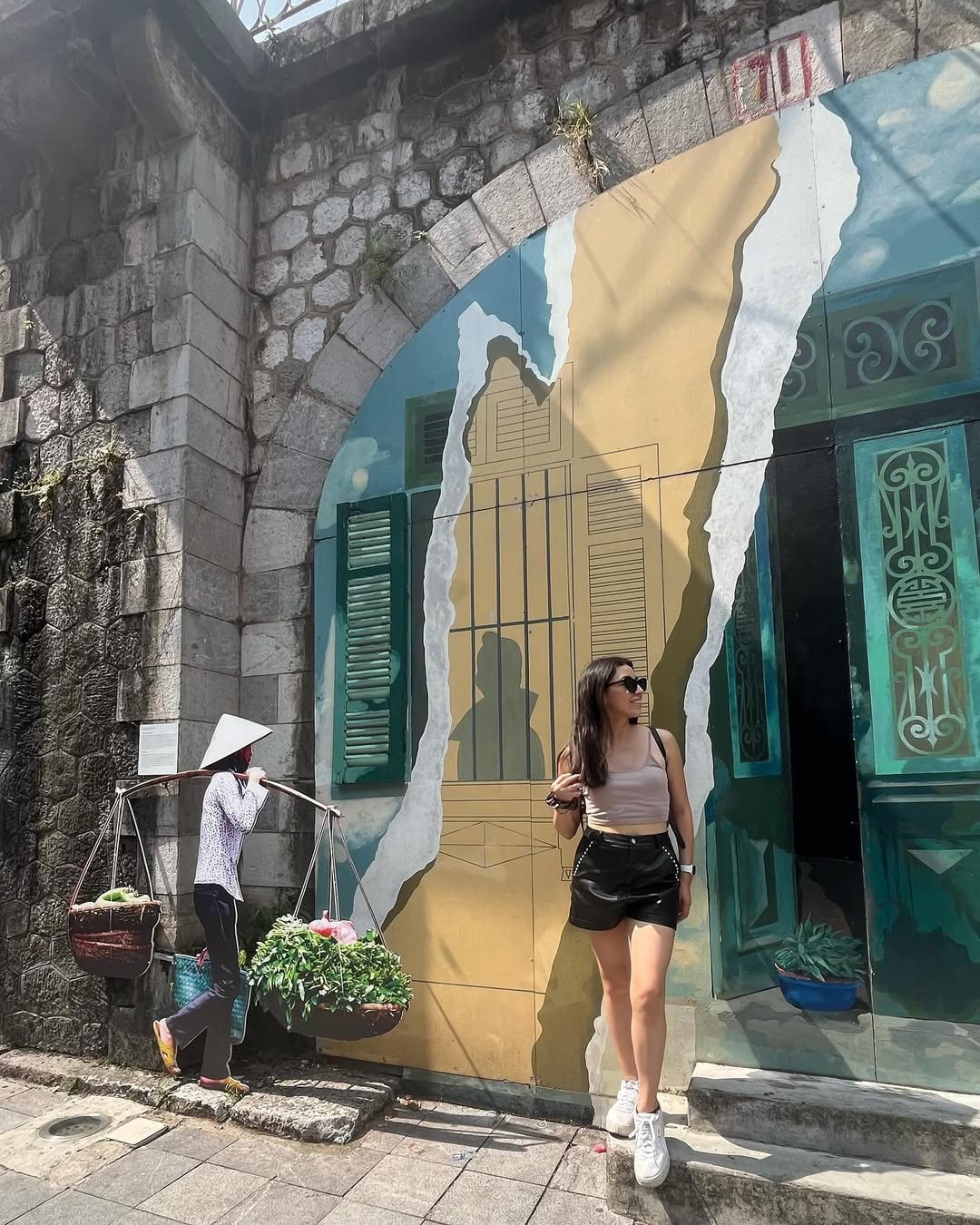Bárbara at a street art mural in Hanoi with conical-hat vendor