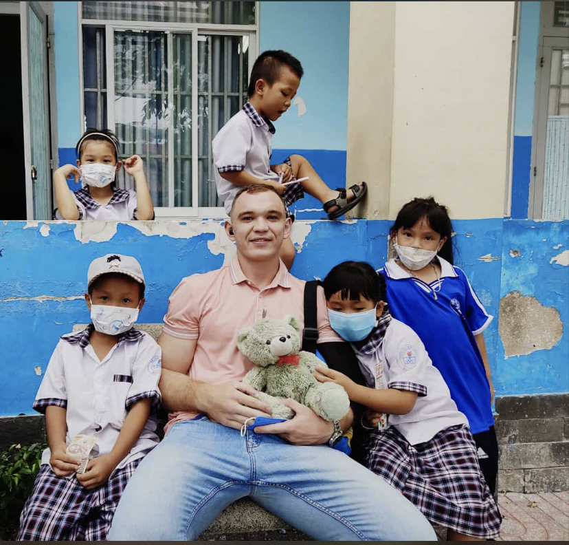 Non-native English teacher sitting with Vietnamese primary school students outside school building